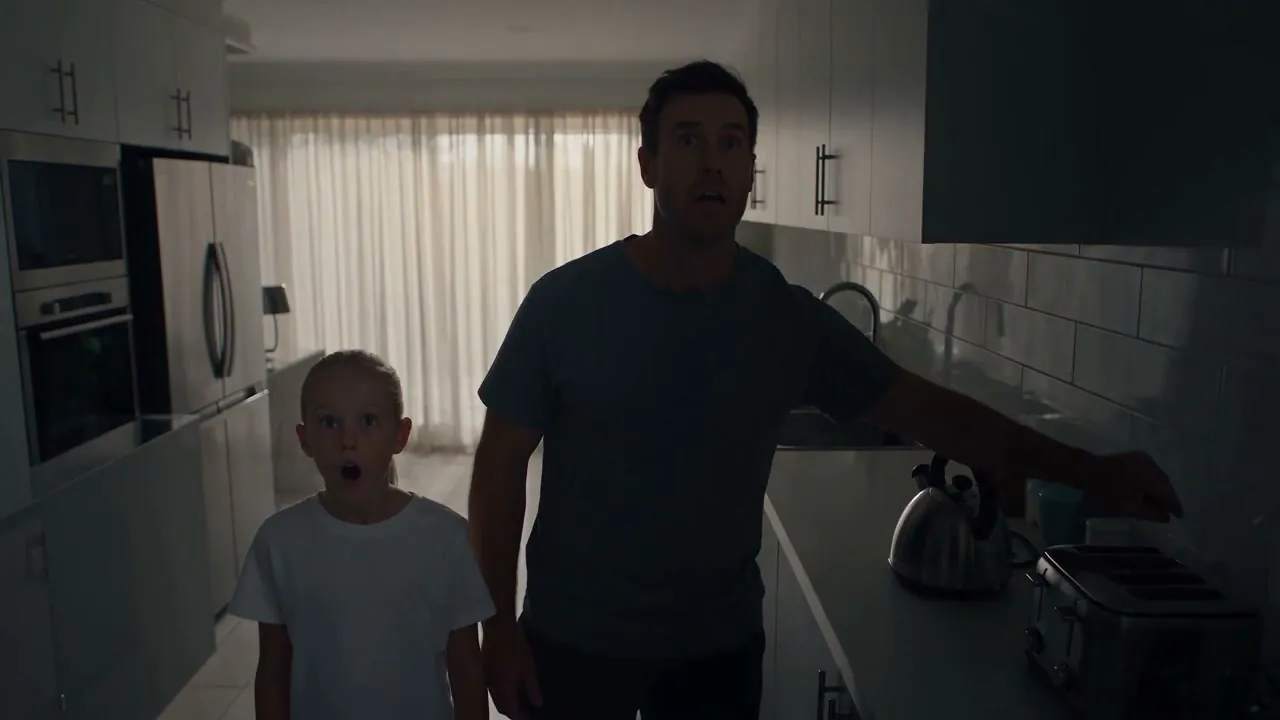 A parent and child walking into a kitchen that has gone dark except for daylight through the window