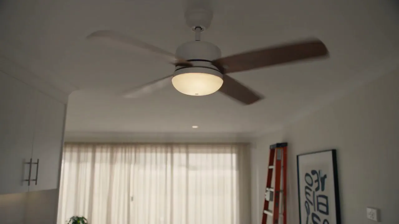 A ceiling fan mid-wobble in a living room, sheer curtains and a step ladder behind it